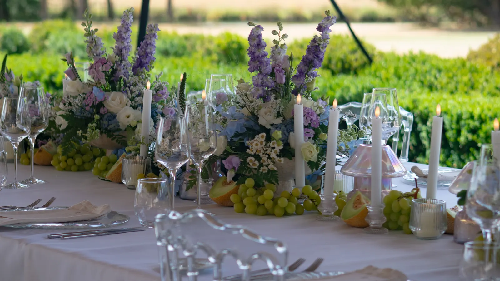 A long table dressed in white linen with lilac and white florals, candles and cut fruit, set out on a lawn beside a clipped hedge