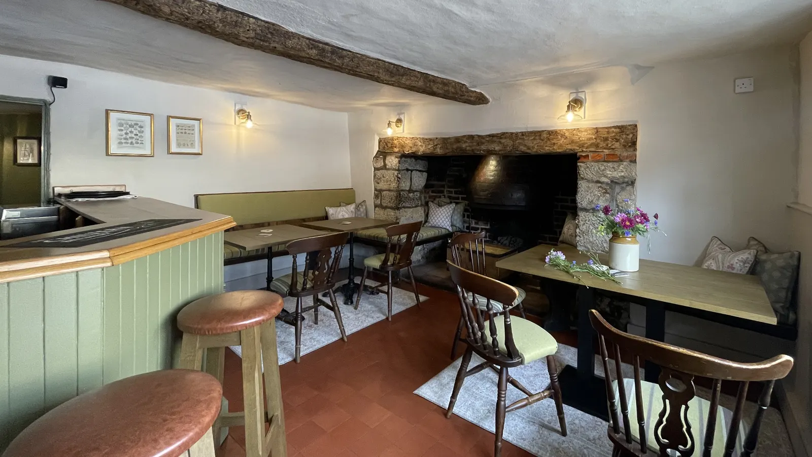 A beamed pub room with a brick inglenook, a green panelled bar, stools and laid tables