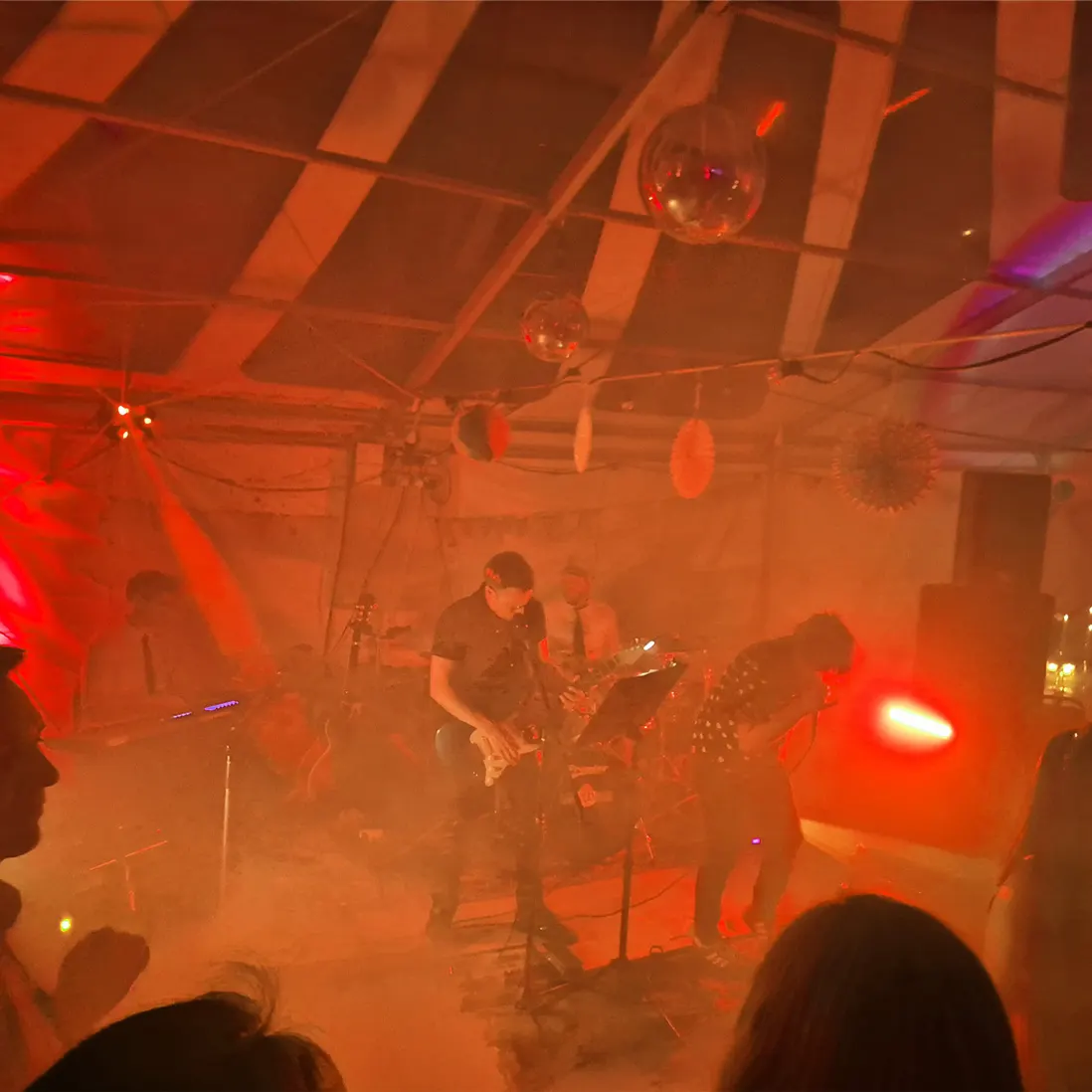 A live band on stage under a marquee, lit in red with haze and mirror balls