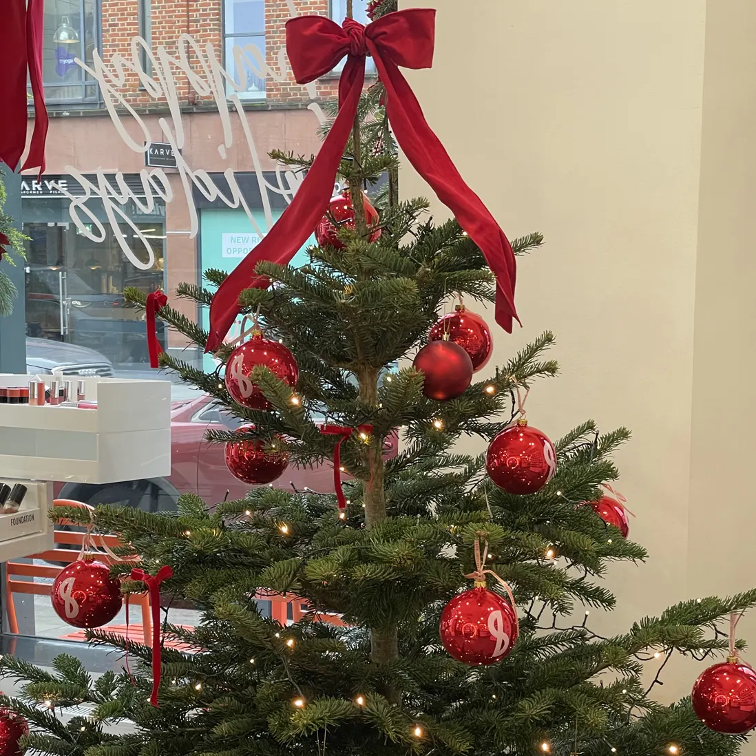 A fir tree hung with red glass baubles and velvet ribbon, lit with fairy lights beside a shop window