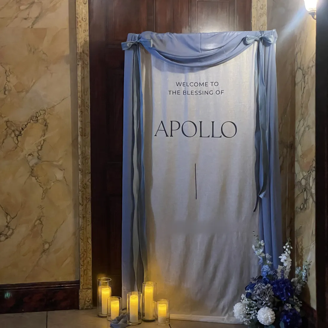 A draped welcome board in dusty blue with candles and urns of blue and white florals at its base