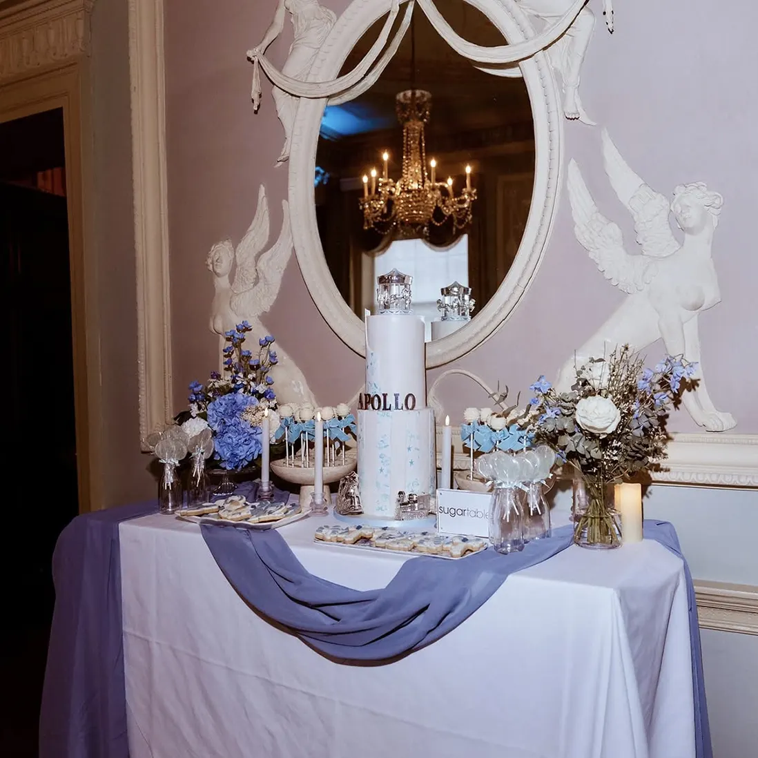 A dessert table dressed in blue and white beneath a gilt mirror, with a tiered cake, florals and draped linen