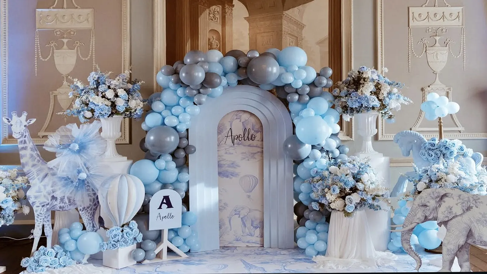 A balloon arch in dusty blue and grey framing an arched toile backdrop, with sculpted animal props and white urns of blue and white florals