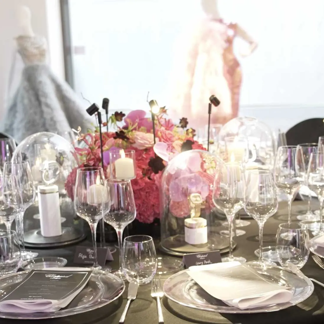 Jewellery on plinths under glass cloches among candles, glassware and pink and red florals on a dressed table