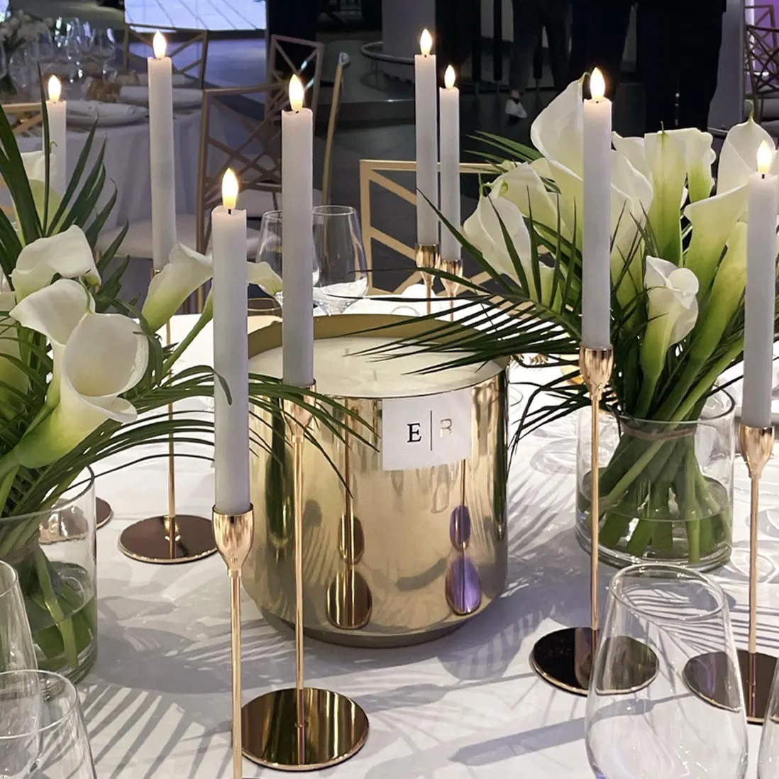White calla lilies in a polished gold vessel between tapered candles and palm leaves