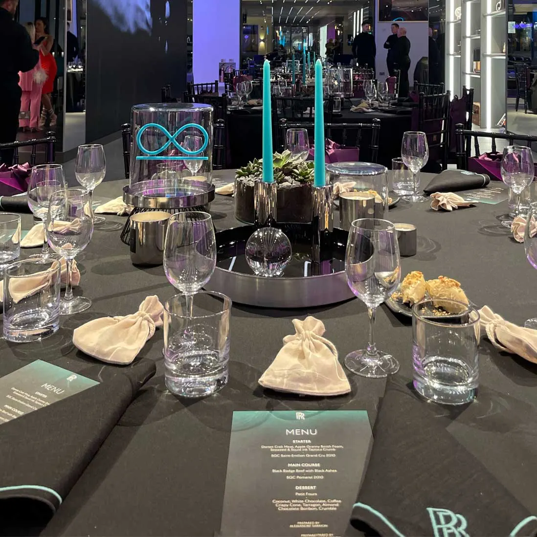 A dark tablescape with teal tapered candles, gold favour pouches and a menu card