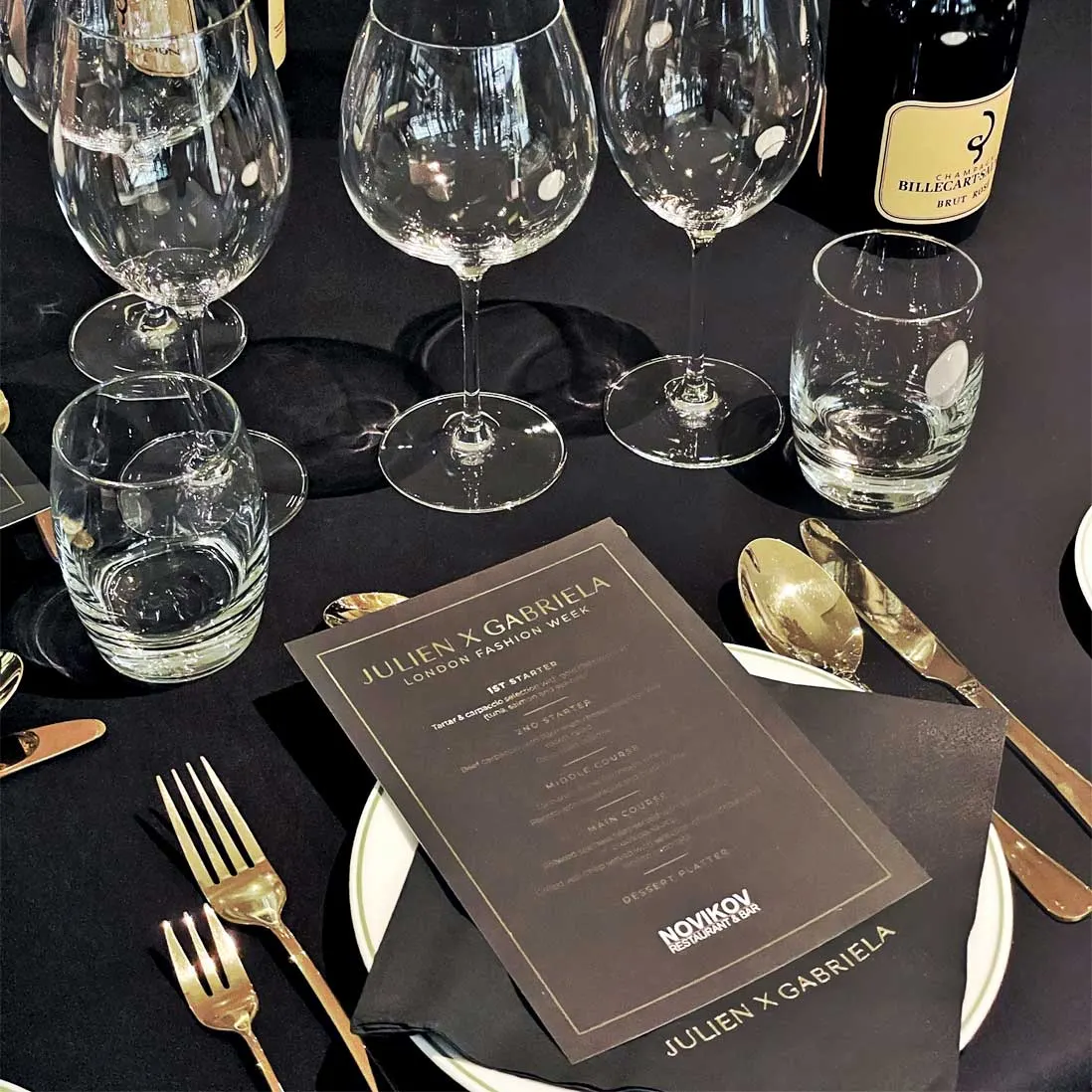 A place setting on black linen with gold cutlery, glassware and a printed menu card