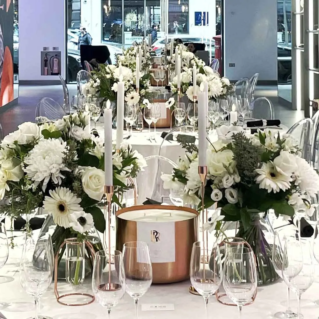 White florals and a copper candle vessel among glassware on a dressed table