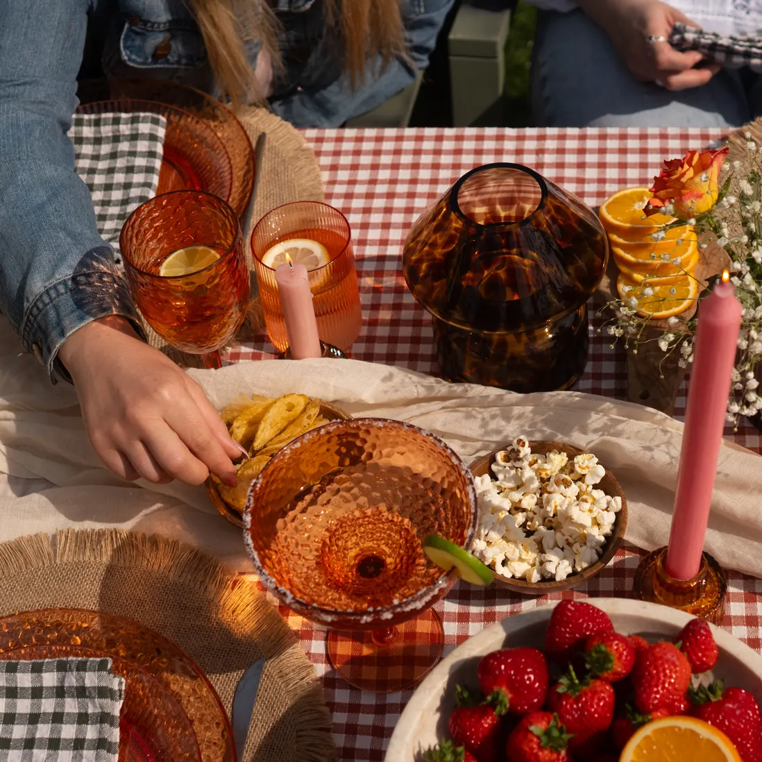 Hands reaching across a gingham-dressed table set with amber glassware, candles and strawberries