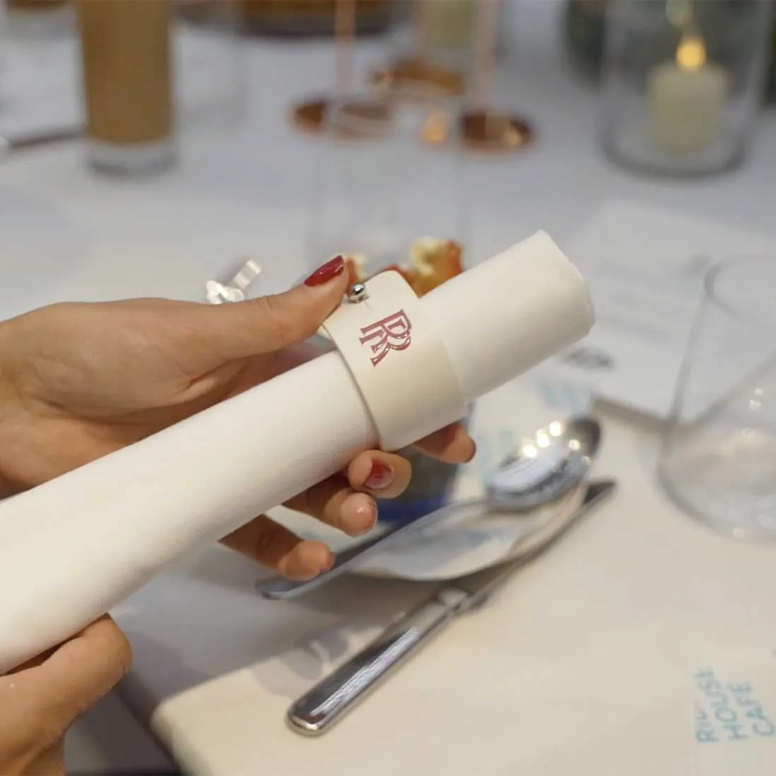 A hand holding a rolled napkin in a monogrammed band above a laid place setting