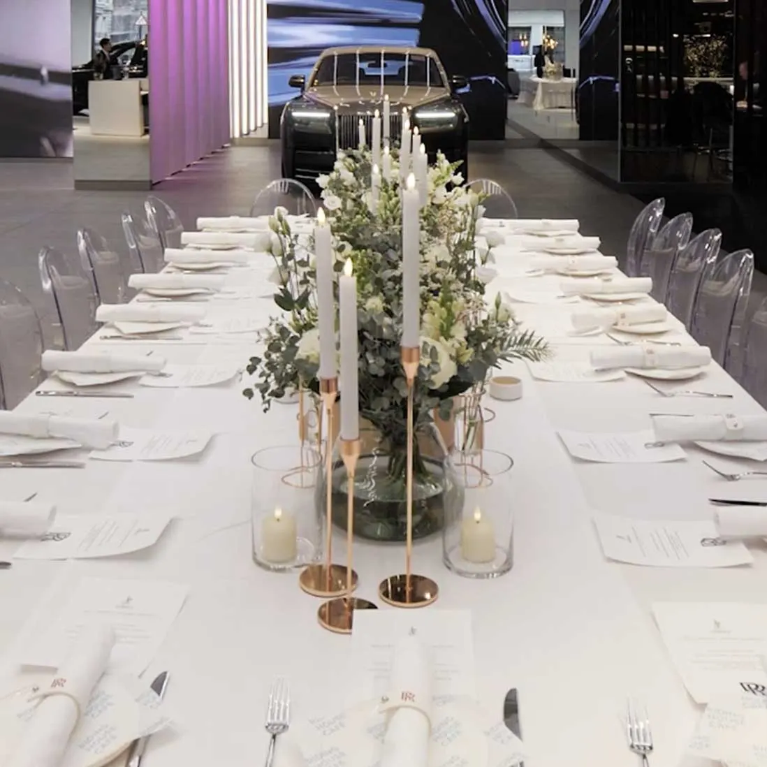 A long breakfast table dressed with foliage, tapered candles and white linen, cars behind