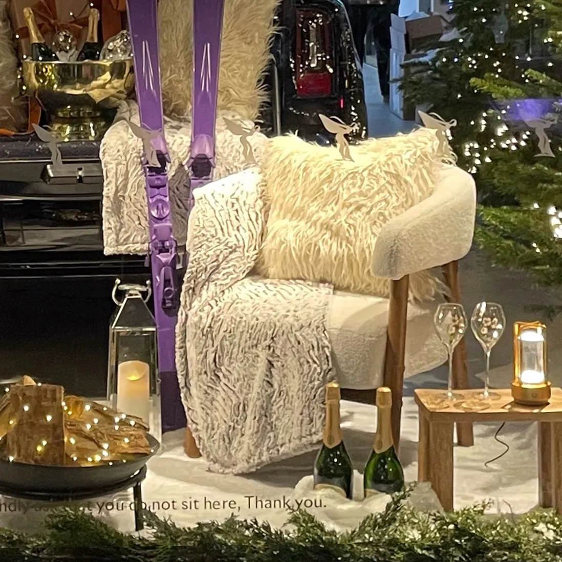 A sheepskin armchair and throw styled beside skis, champagne glasses and a lit lantern in artificial snow