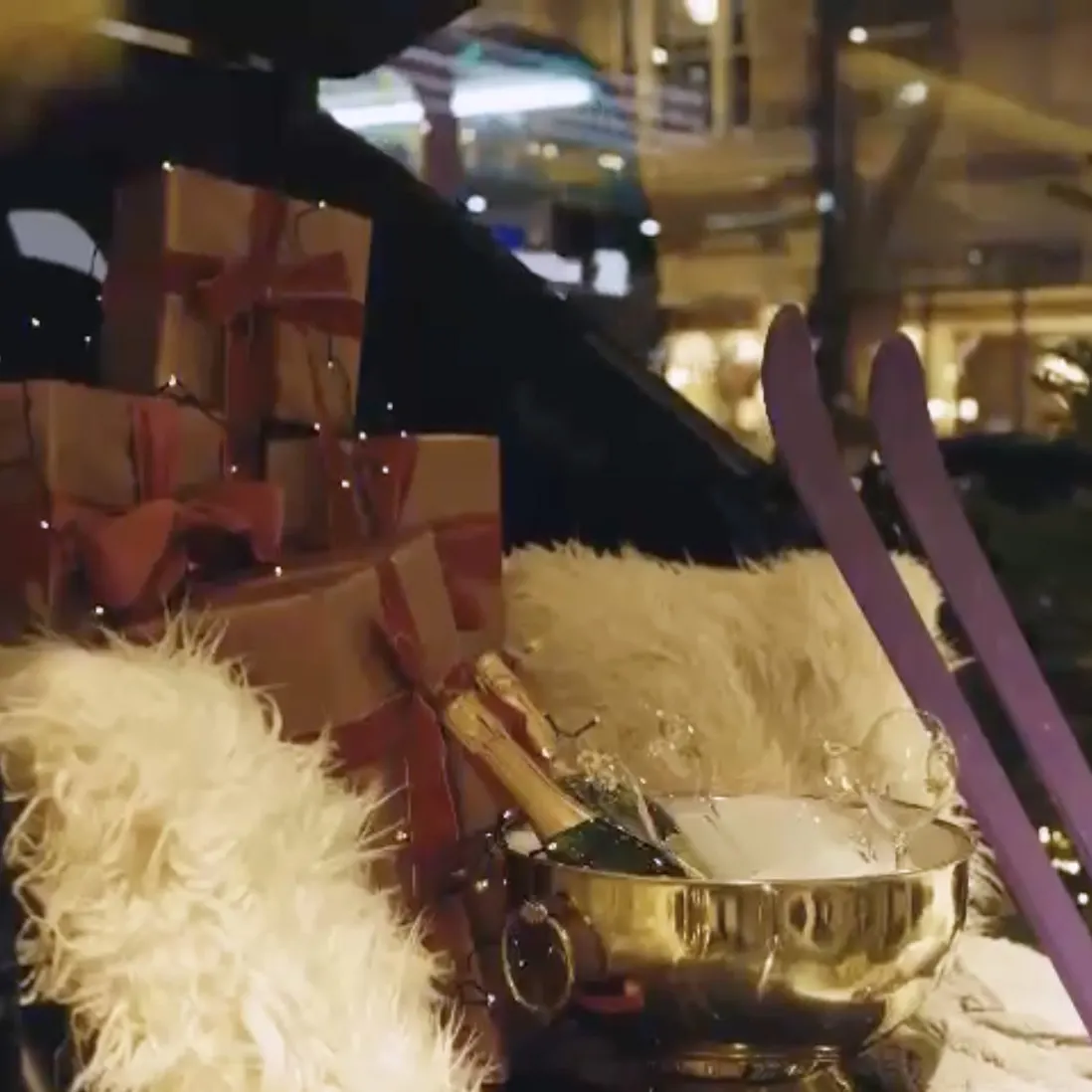 Gift boxes, sheepskin and a champagne bottle in a gold ice bowl, lit beside skis in a winter window