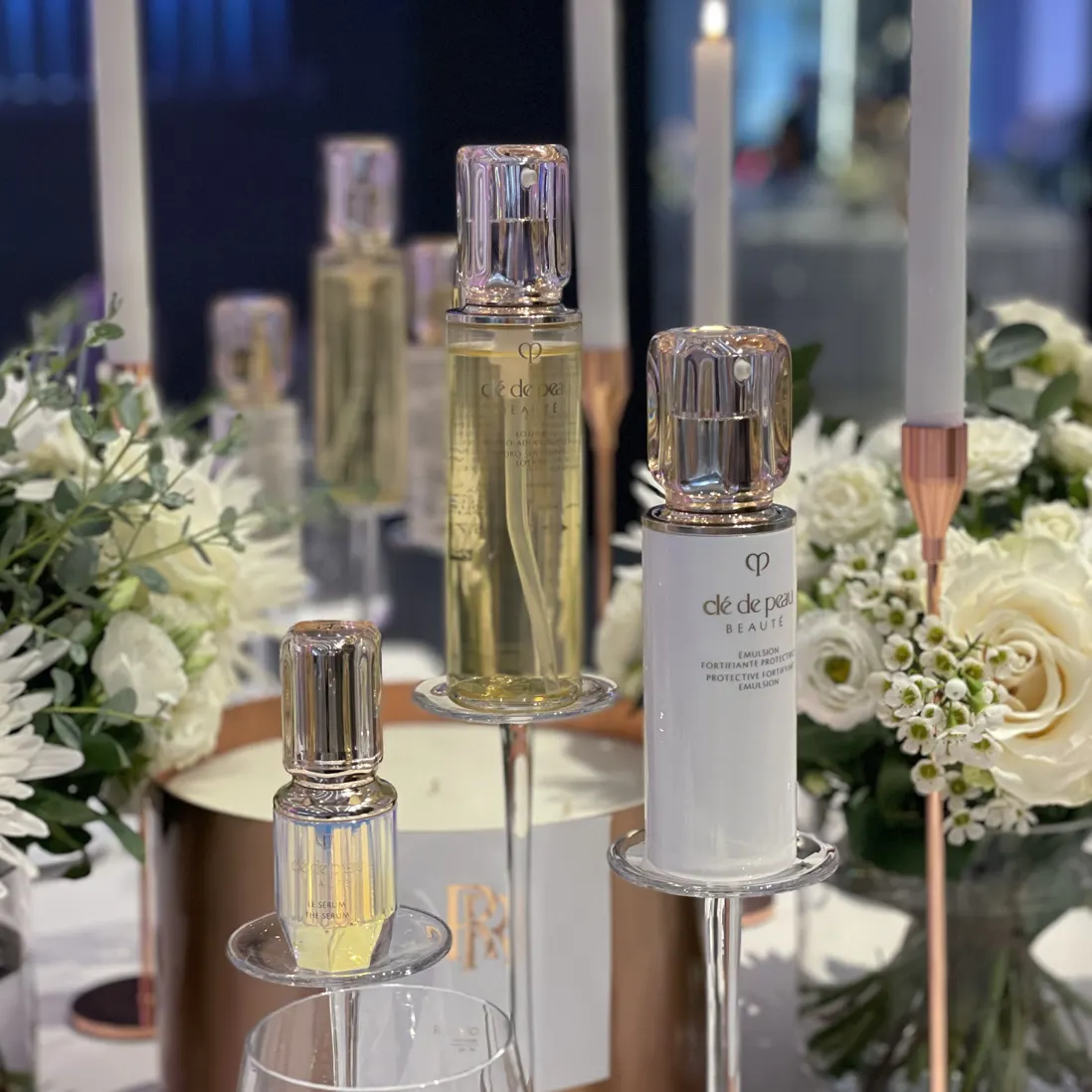 Skincare bottles on lit plinths between tapered candles and white florals