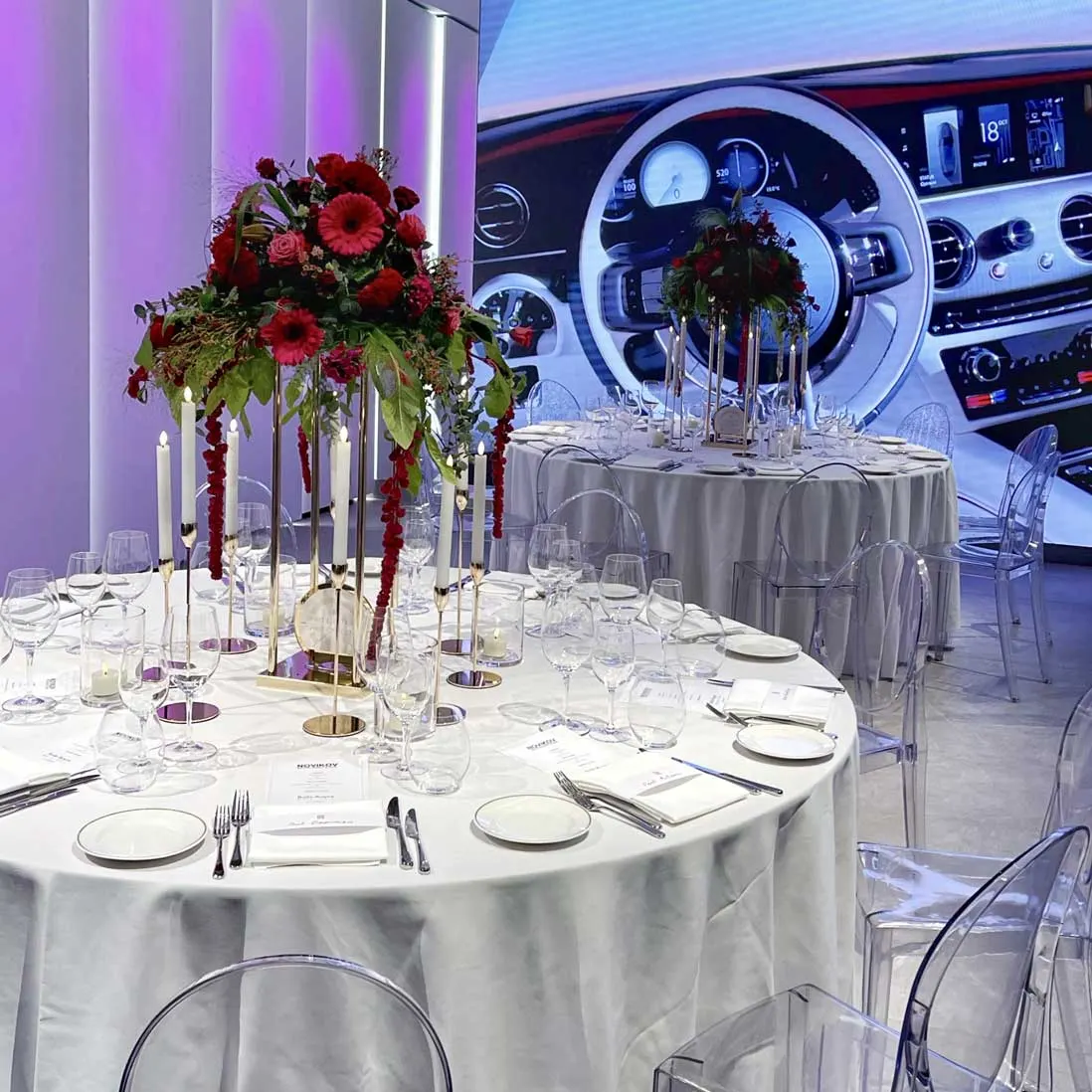 A round table with tall red floral centrepieces and candles set beside a car