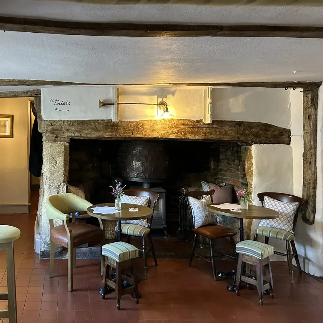 A beamed room with a stone fireplace, bar stools and small tables