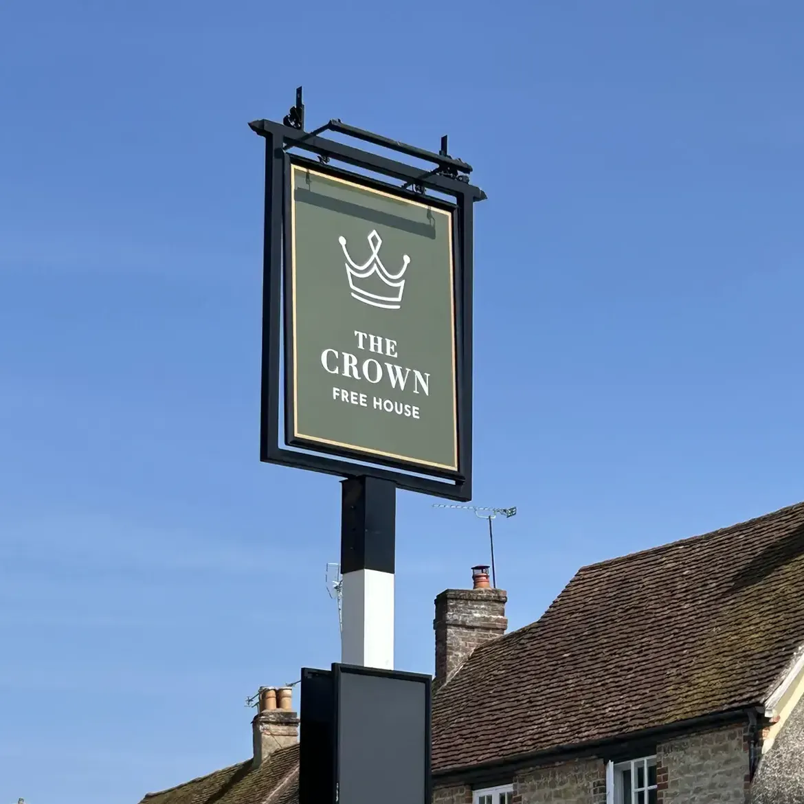 A new hanging pub sign in deep green carrying a line-drawn crown, above the village rooftops